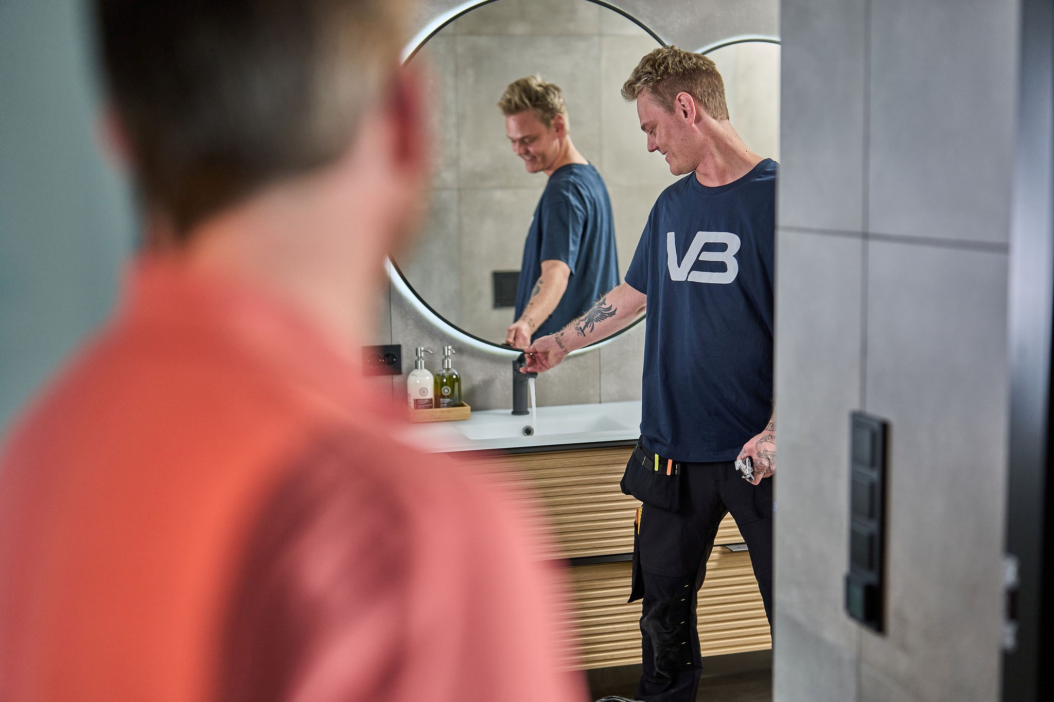 VB-rørlegger tester kran på bad med en annen mann i forgrunnen som ser på.